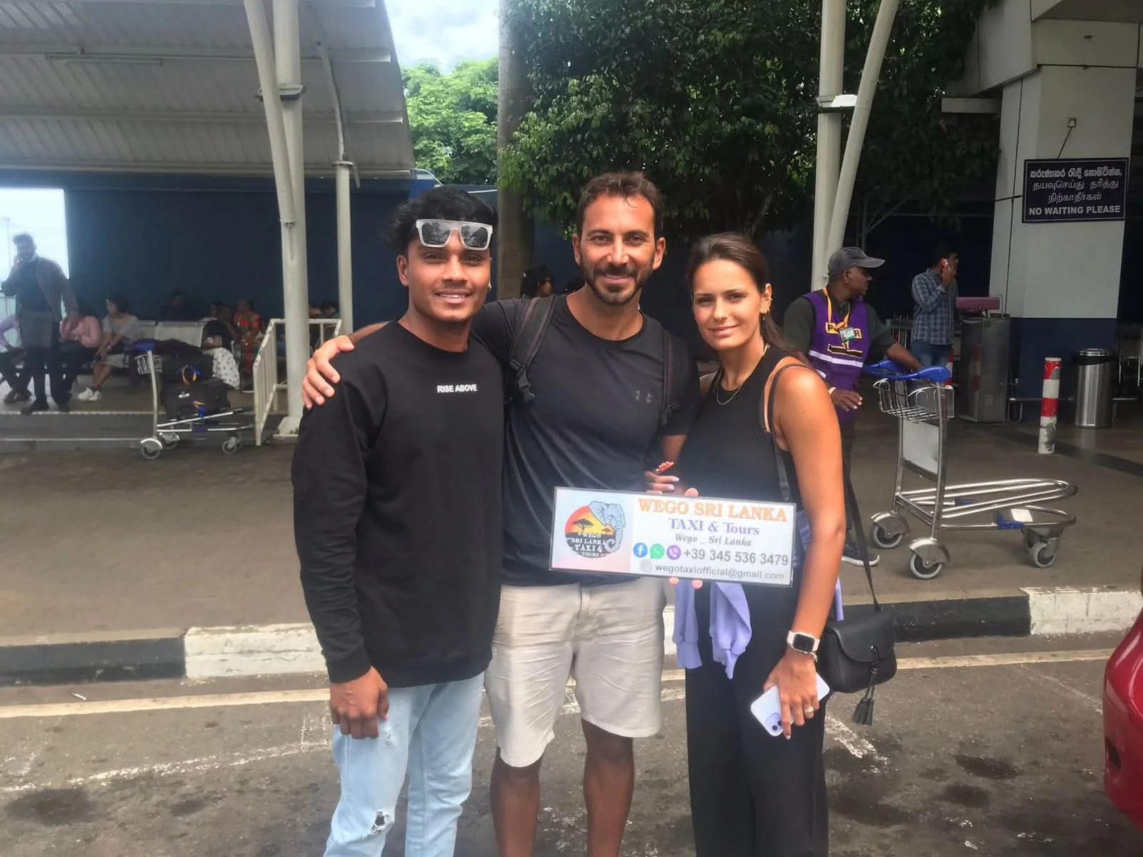 Happy tourists with Sri Lankan driver at airport departure - International couple with professional guide holding Wego Taxi Tours sign after successful tour completion